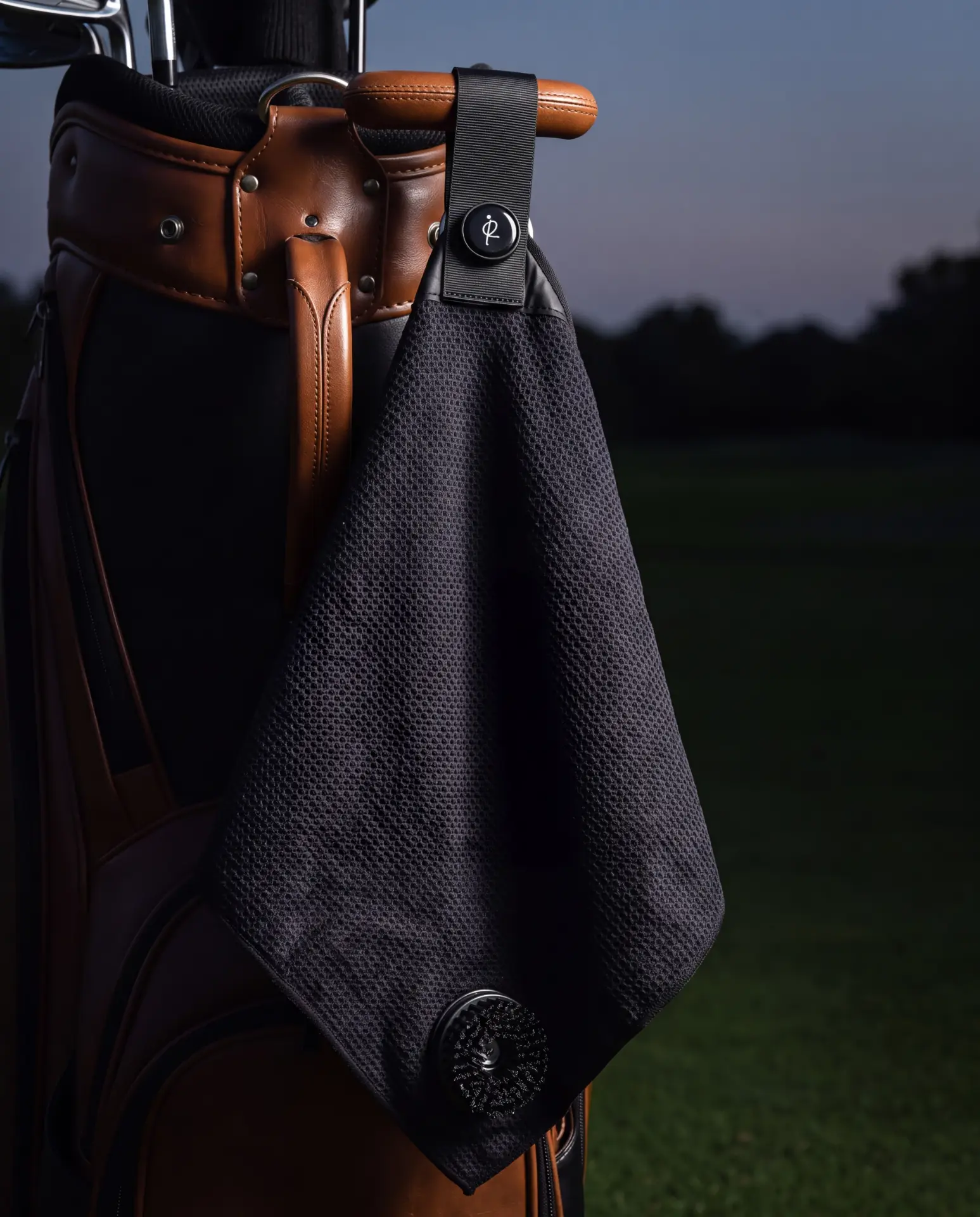 A black golf towel hanging from a golf bag at dusk, held in place by its magnetic strap clipped over the bag rail