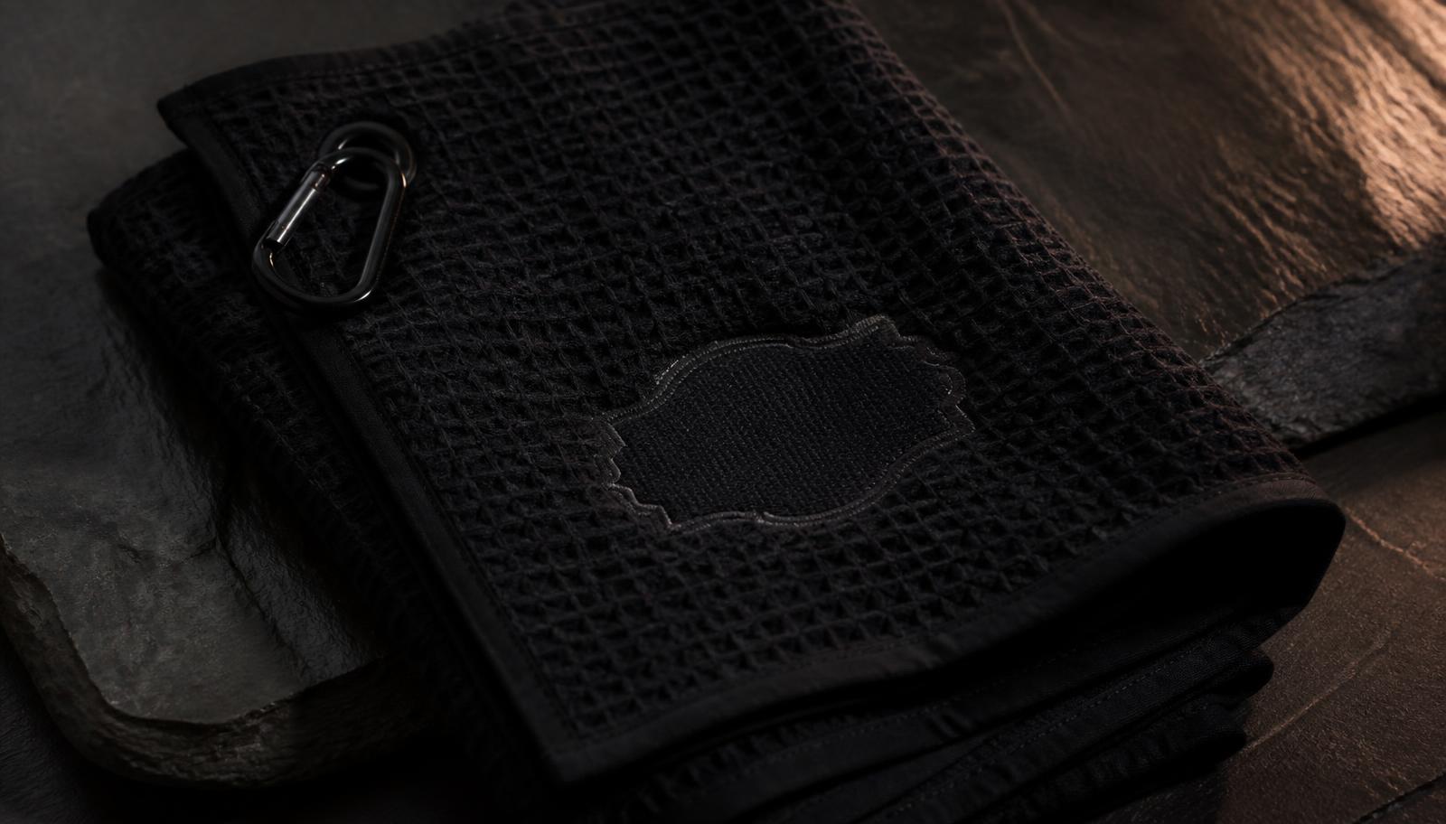A dark folded towel with embroidered monogram stitching, lit from the side with warm low-key light on a slate background
