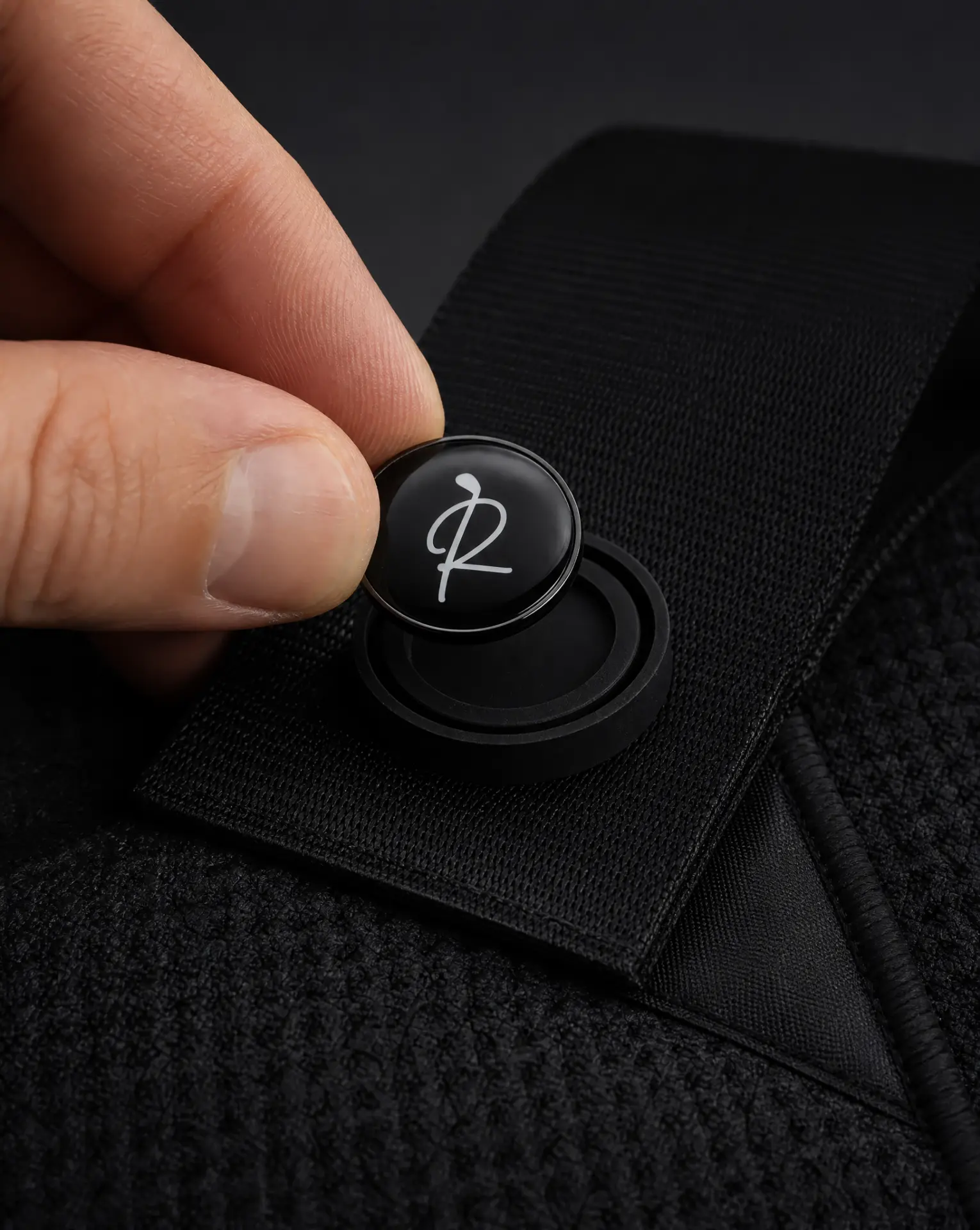 A hand lifting the round magnetic ball marker off the strap of a black Ryft golf towel