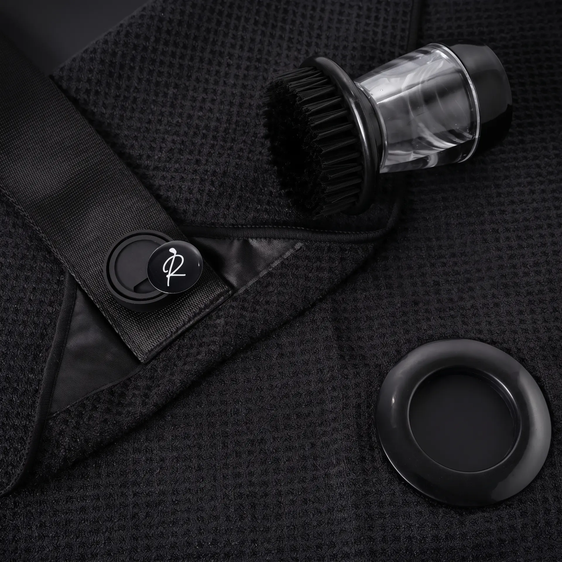 Flat-lay of a black waffle-weave golf towel laid open with its magnetic strap, ball marker disc and brush tool arranged beside it on a dark surface