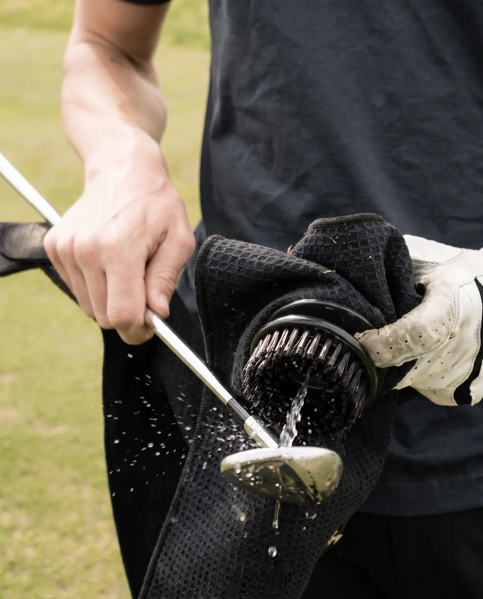 A golfer wiping an iron clubhead clean with a black waffle-weave towel and brush after a shot